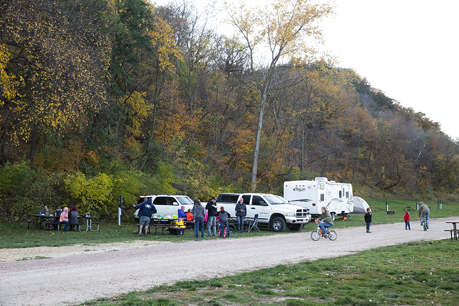 Groups of people picnicking with two people riding bikes while children look on in the background. Two SUVs and a RV are in the background as well with fall foliage beyond.